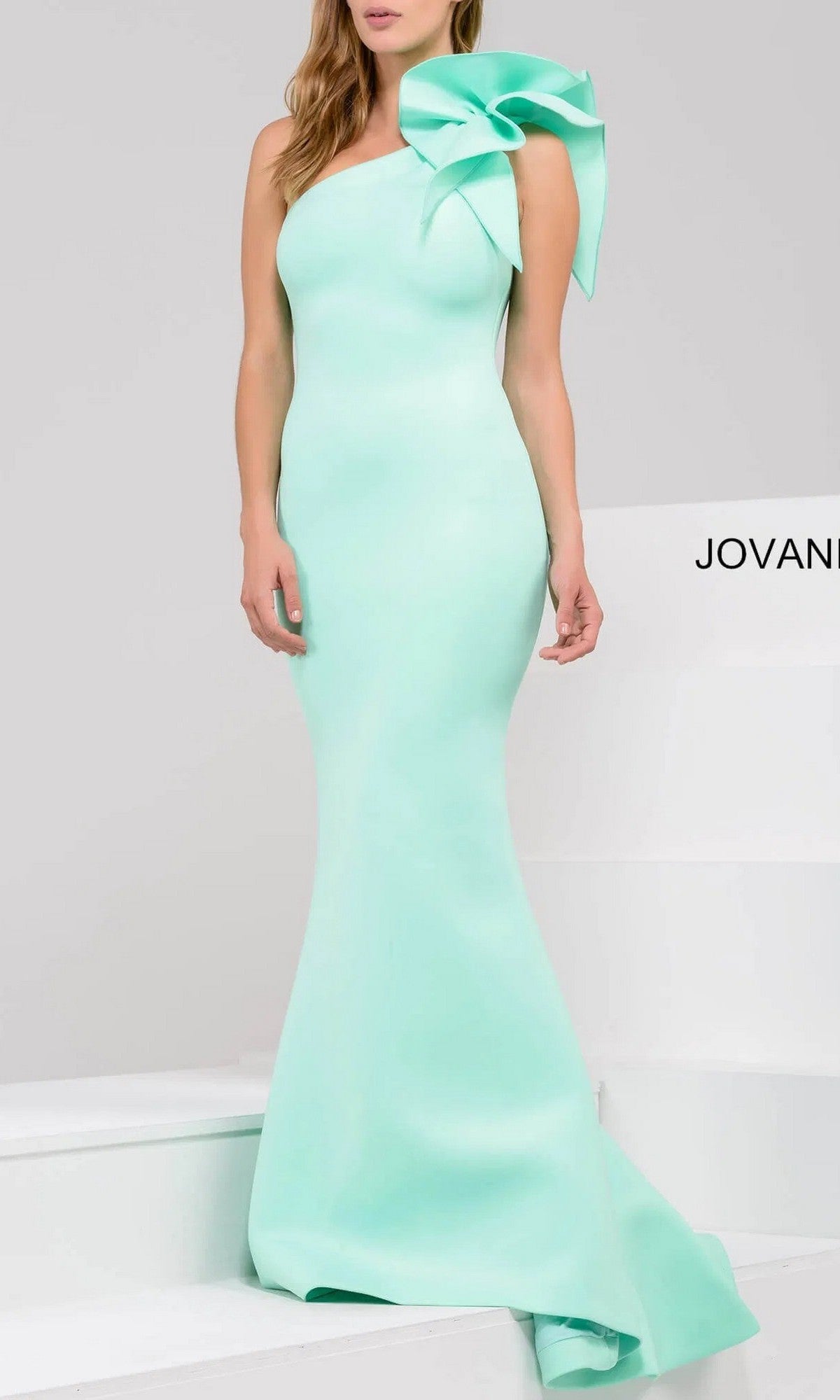 A woman stands on white steps wearing the Jovani 32602, a fitted mint green one-shoulder mermaid prom gown featuring a large bow detail on the shoulder.